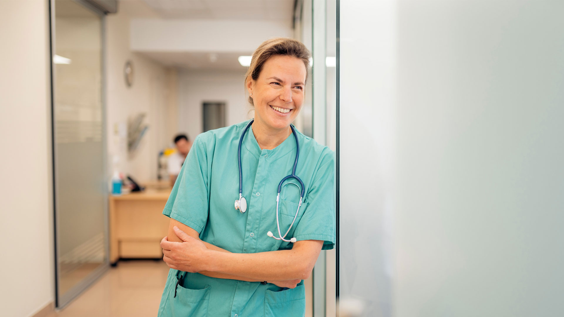 woman doctor wearing teal scrubs and a stethoscope around her neck leaning agains a wall casually smiling