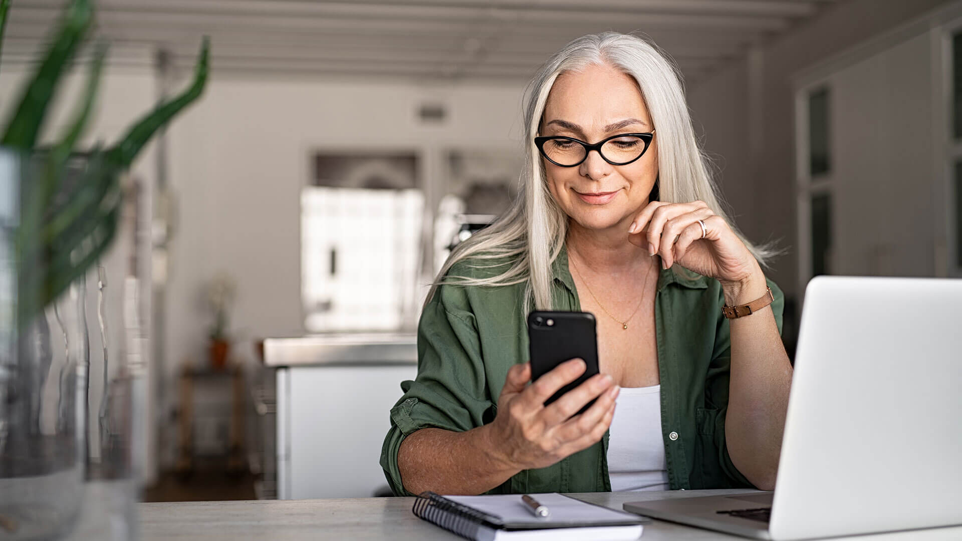 middle aged woman wearing glasses using mobile phone at with laptop on desk